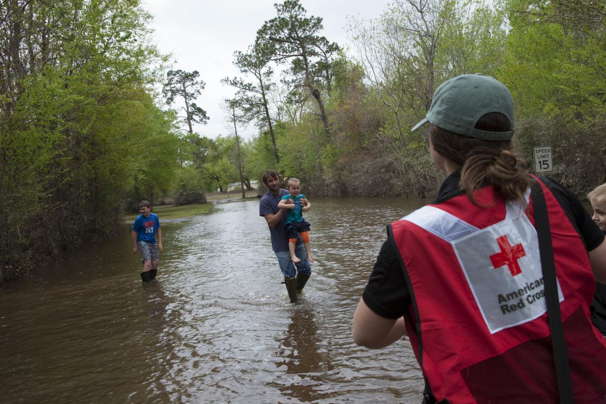 March 23, 2016. Deweyville, Texas. In Deweyville Texas, many roads are still underwater making access to homes nearly impossible. The Parkers have to cross the flooding waters in order to begin clean up in their home. Their 2 year old daughter, Aubrey, slipped while walking across and her 12 year old brother, Nathan, jumped into action and grabbed her from the water. Red Cross volunteer, Karli Epstein, assists the parents as they make their way across the road. Carla Parker takes her scared daughter in her arms and back to safety. Photo by Danuta Otfinowski/American Red Cross